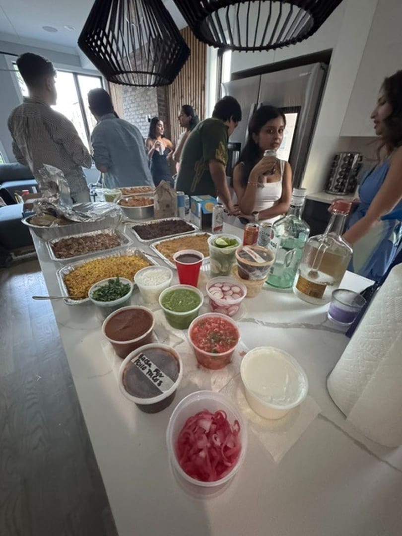 A buffet spread of Mexican food and condiments on a kitchen island with people mingling.