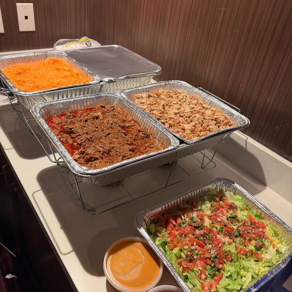 A buffet spread with foil pans of shredded meat, rice, lettuce, pico de gallo, and sauces.