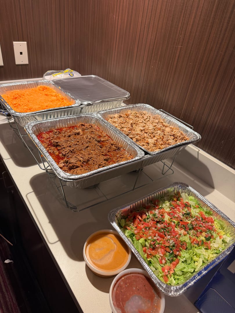 A buffet spread with foil pans of shredded meat, rice, lettuce, pico de gallo, and sauces.