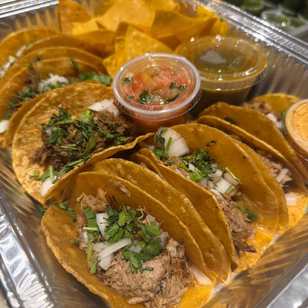 A foil container filled with street tacos, tortilla chips, salsa, and green sauce.