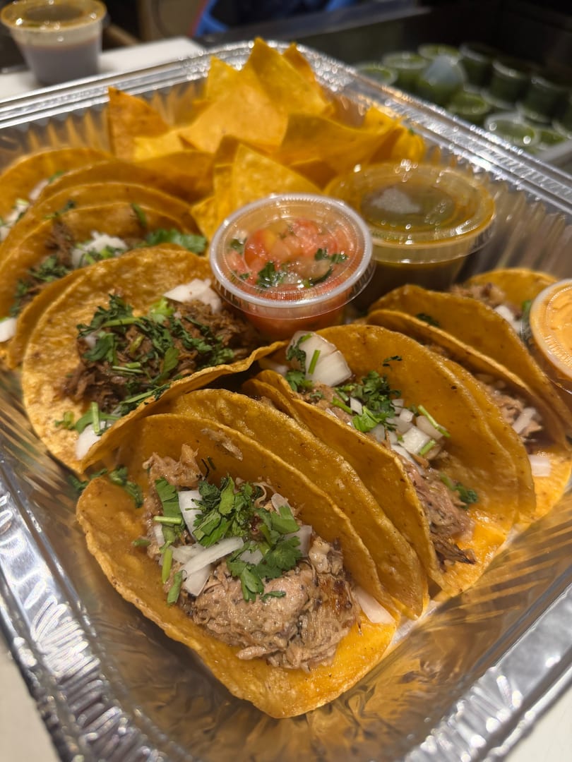 A foil container filled with street tacos, tortilla chips, salsa, and green sauce.